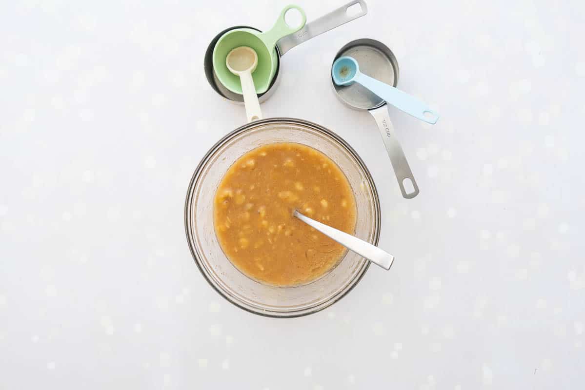 A bowl of banana bread wet ingredients on a bench top surrounded by mixing spoons and cups.  