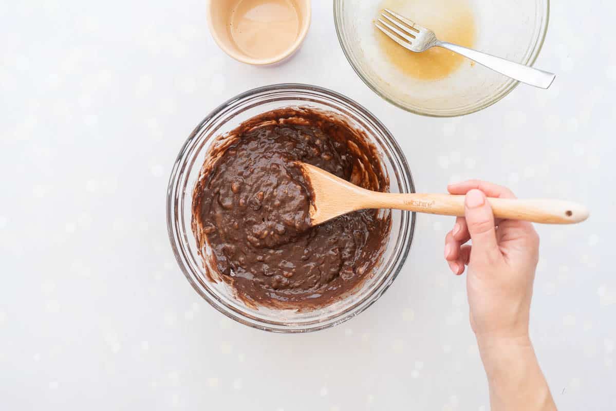 Chocolate banana bread batter in a glass mixing bowl, being stirred with a wooden spoon.