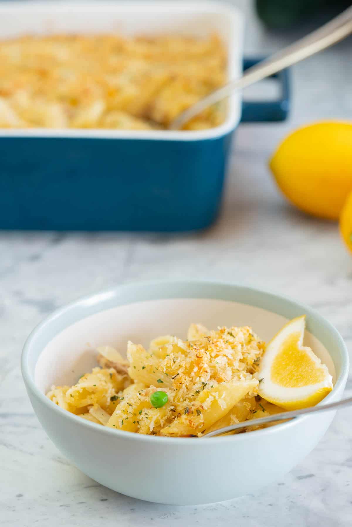 A bowl of creamy penne pasta on a grey marble bench top with a dark blue baking dish and lemons in the background.