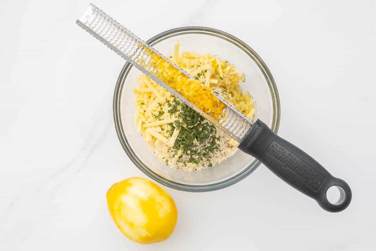 A glass mixing bowl containg bread crumbs, lemon zest parsley and grated cheese, a lemon zester is resting on the bowl.