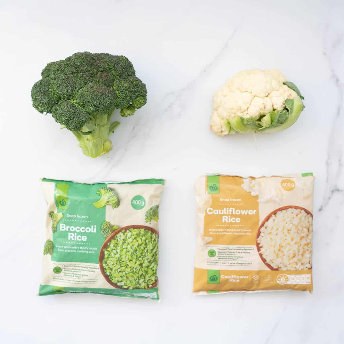 A head of broccoli next to a pack of frozen broccoli rice, a head of cauliflower next to a bag of frozen broccoli rice.