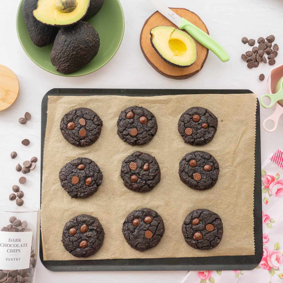 A tray of baked avocado cookies on a cookie tray next to a bowl of avocados and jar of chocolate drops,