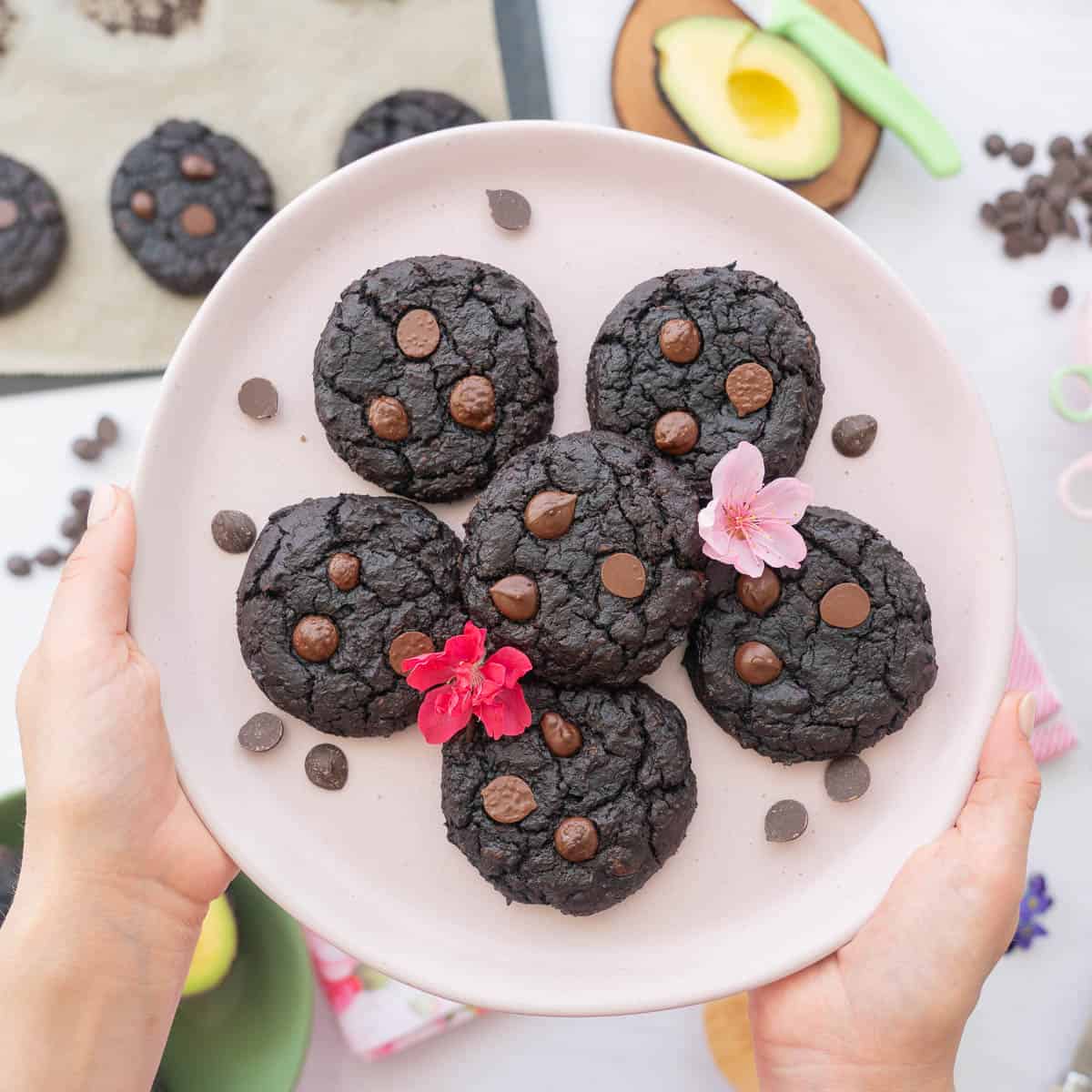 Six chocolate cookies on a pale pink plate scattered with edible flowers.