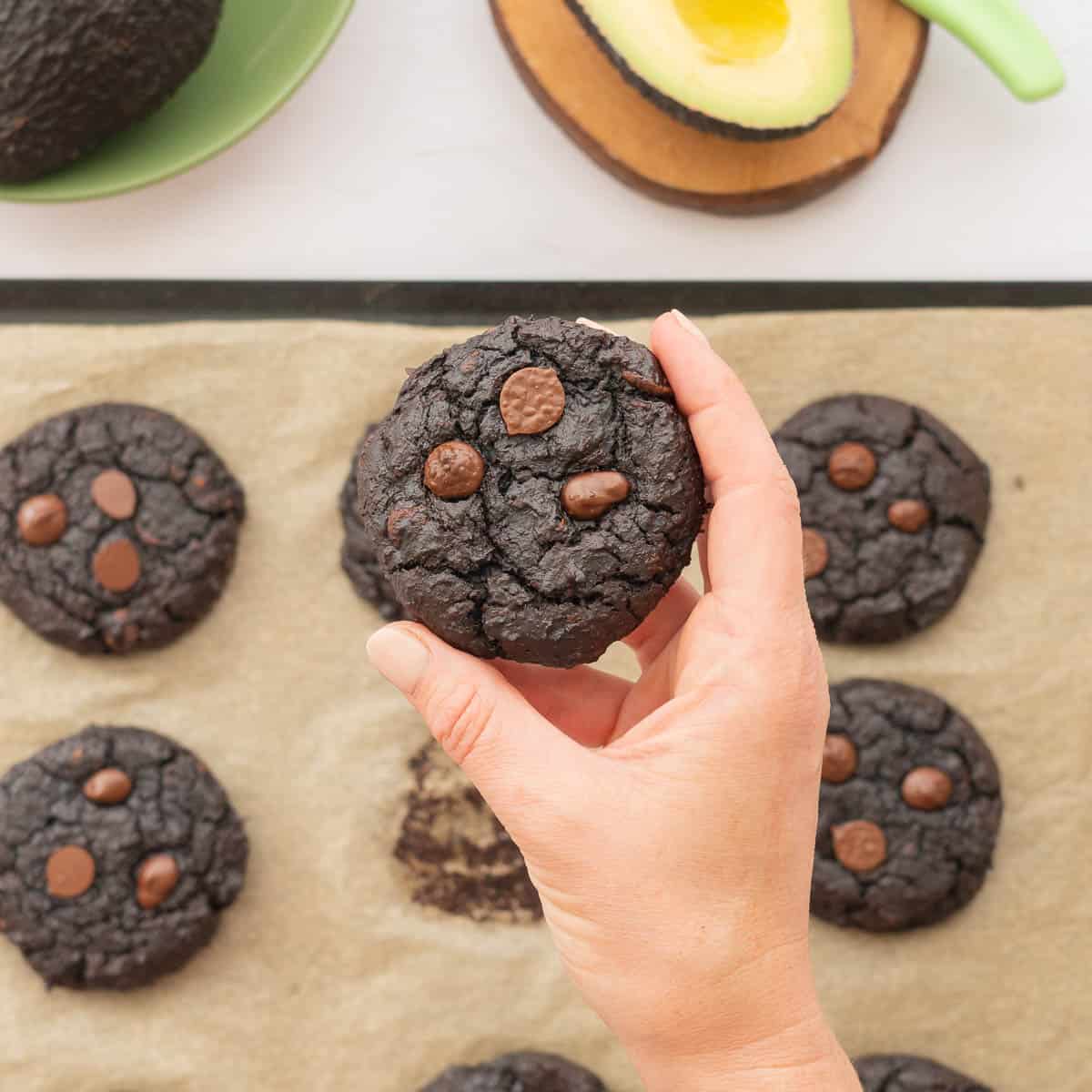 A chocolate cookie being held above a baking tray and bowl of halved avocados.