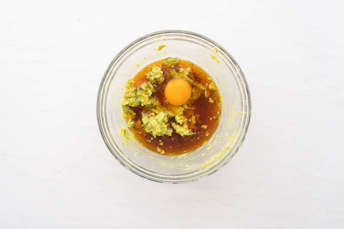 A glass mixing bowl with mashed avocado, raw egg and maple syrup.