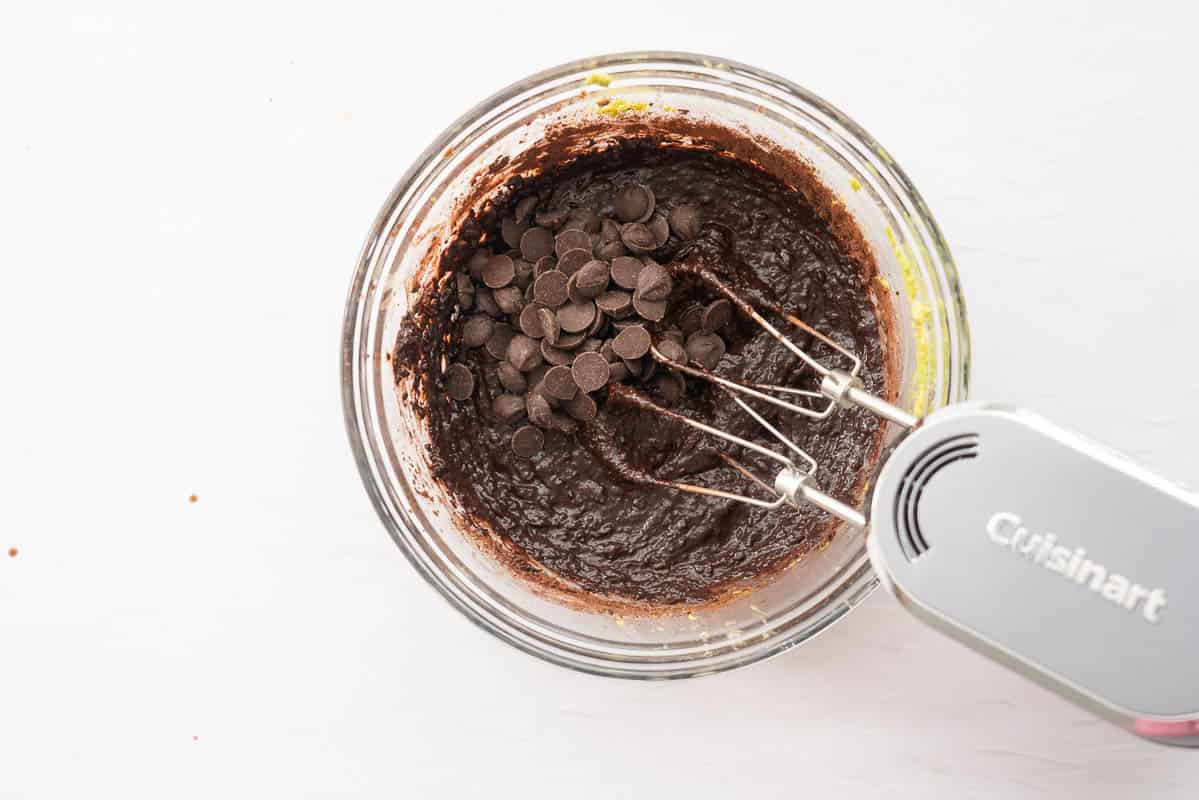 Chocolate avocado cookie batter and dark chocolate drops in a glass mixing bowl with a hand held electric beater.