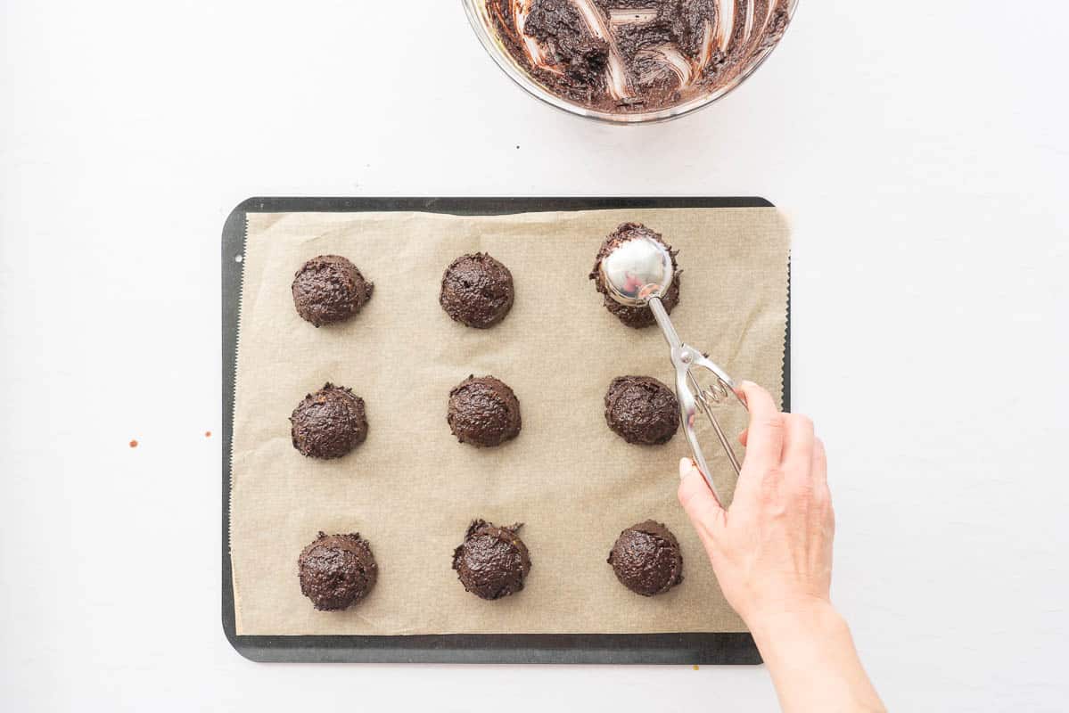 Chocolate cookie batter being scooped onto a baking paper lined tray.