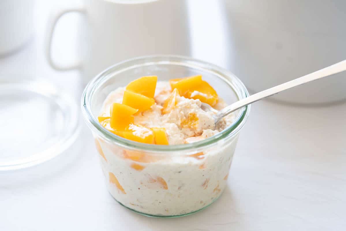 A parfait spoon digging into a glass jar of creamy oats topped with diced peaches.