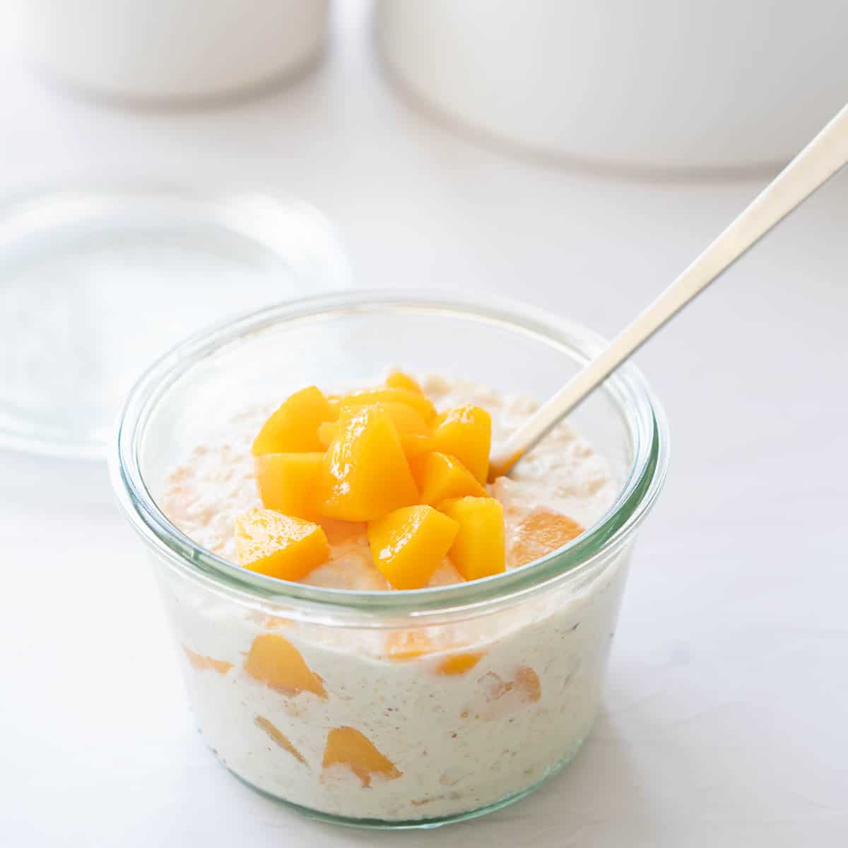 A glass jar of creamy white oats topped with diced peaches, with a long parfait spoon.