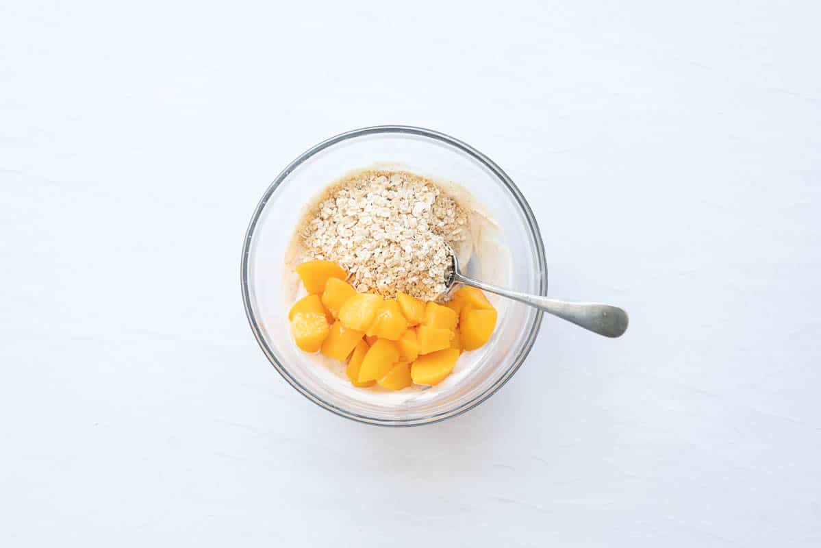 Flavoured yoghurt, diced peaches and rolled oats in a small mixing bowl.