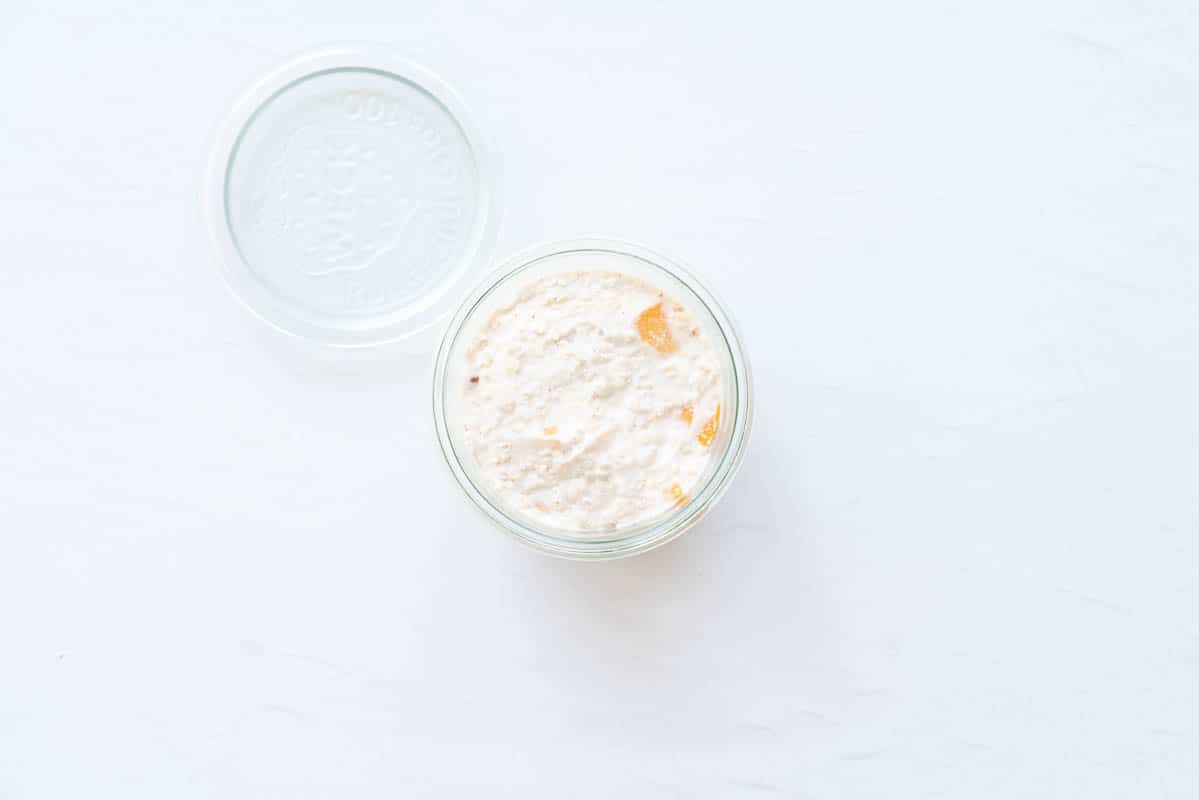 Creamy oats with small pieces of orange peach in a glass jar next to its lid. 