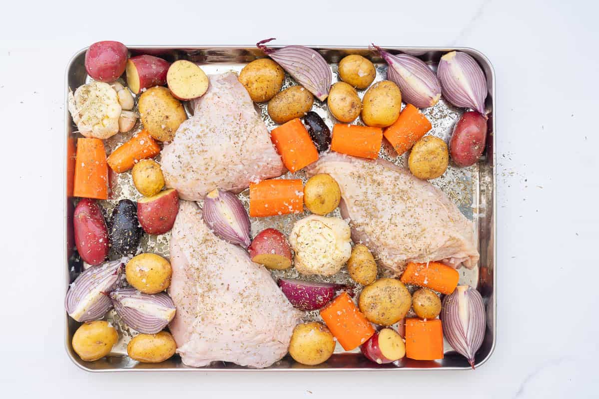 Vegetables and three chicken breasts drizzled in oil and dried herbs in a large stainless steel tray.