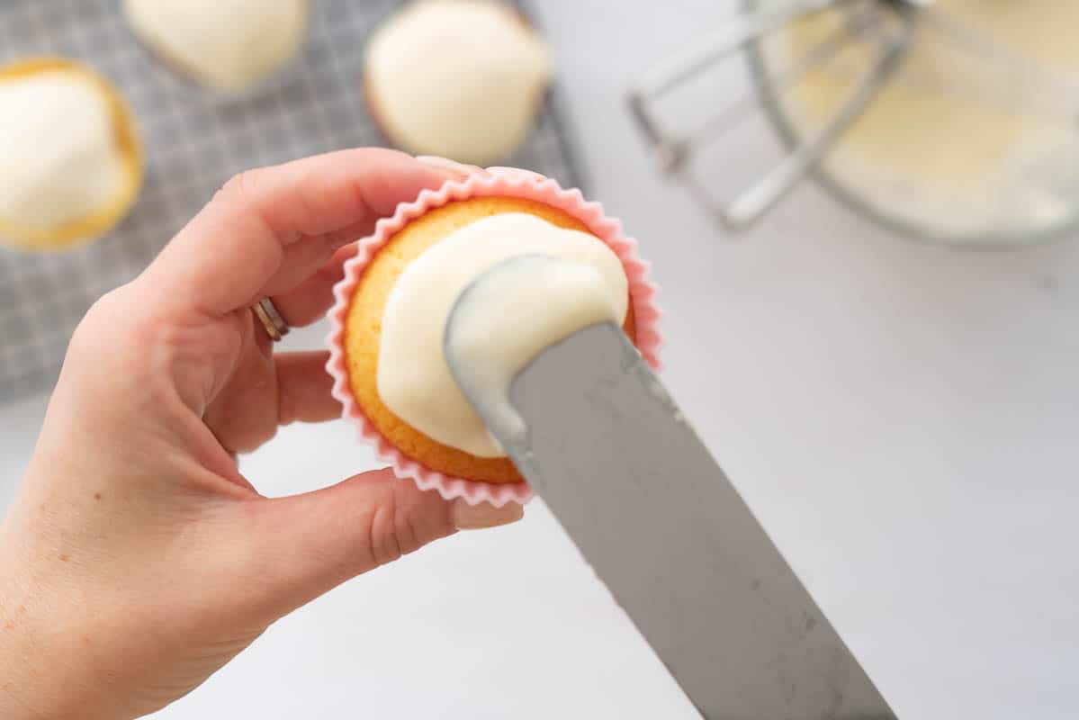 Vanilla glaze being spread onto a cupcake with a cake small baking spatula. 