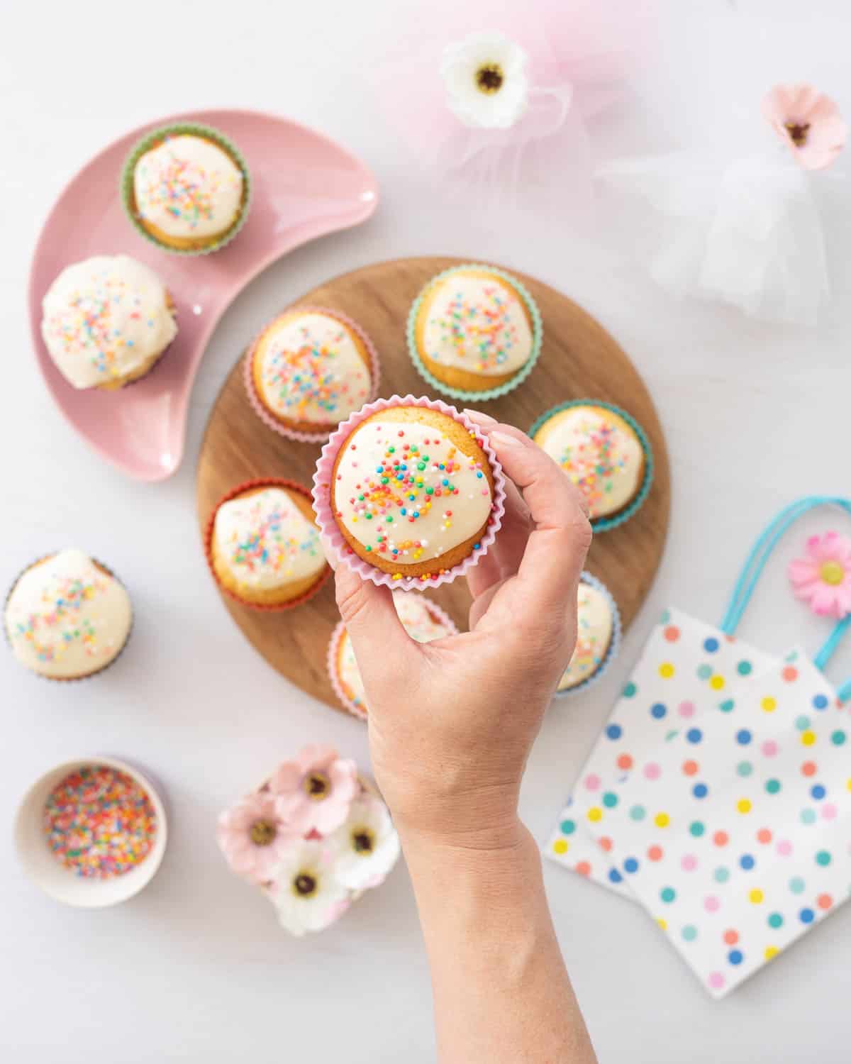 A cupcake in a pink cupcake case decorated with white glaze and rainbow sprinkles being held above a wooden board of more cupcakes. 