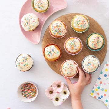 A child's hand reaching for cupcakes decorated with white frosting and sprinkles on a wooden chopping board.