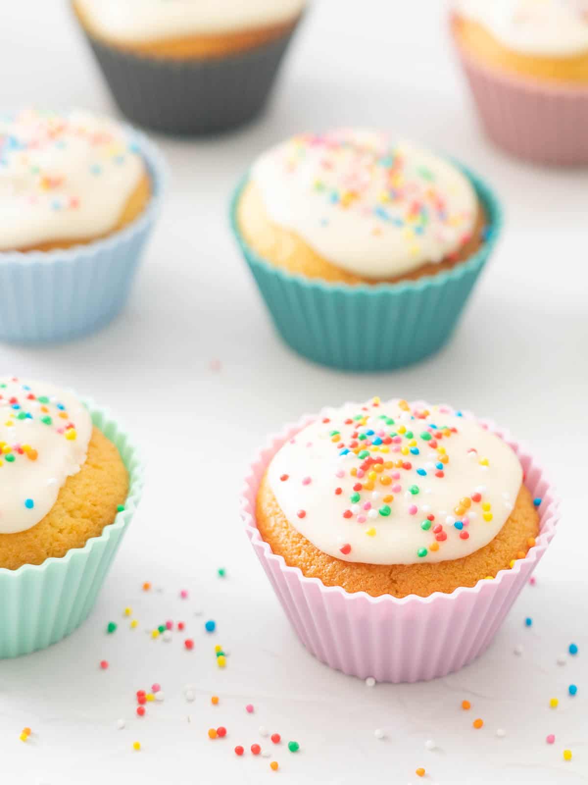 Cupcakes decorated with white glaze and rainbow sprinkles on a white bench top.