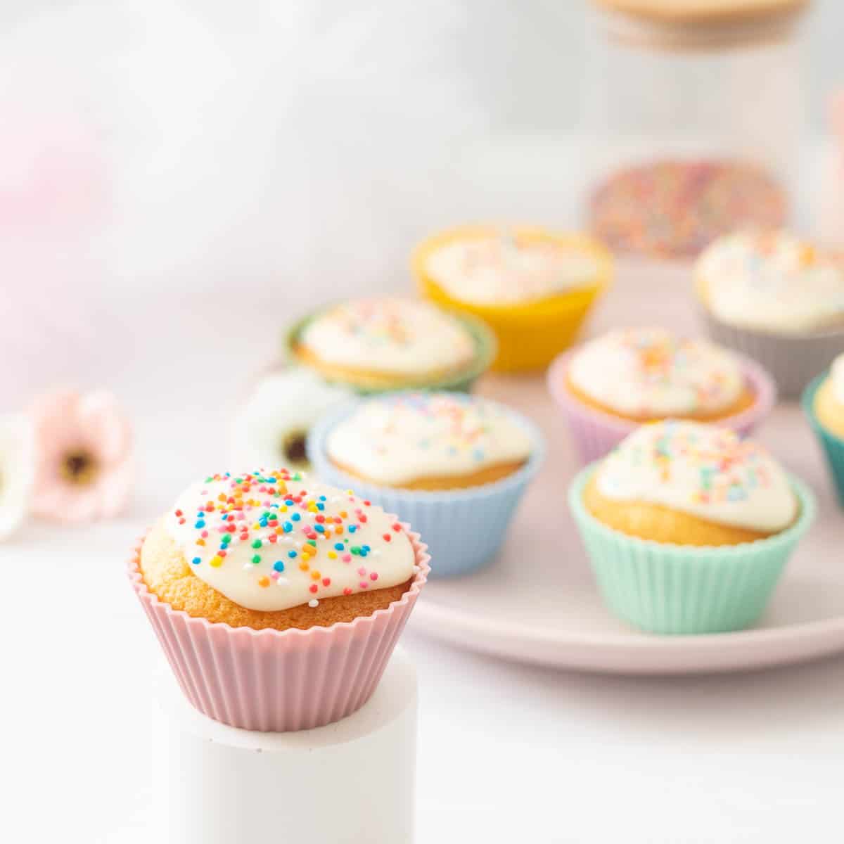 Vanilla cupcakes decorated with rainbow sprinkles displayed on white and pink plates.