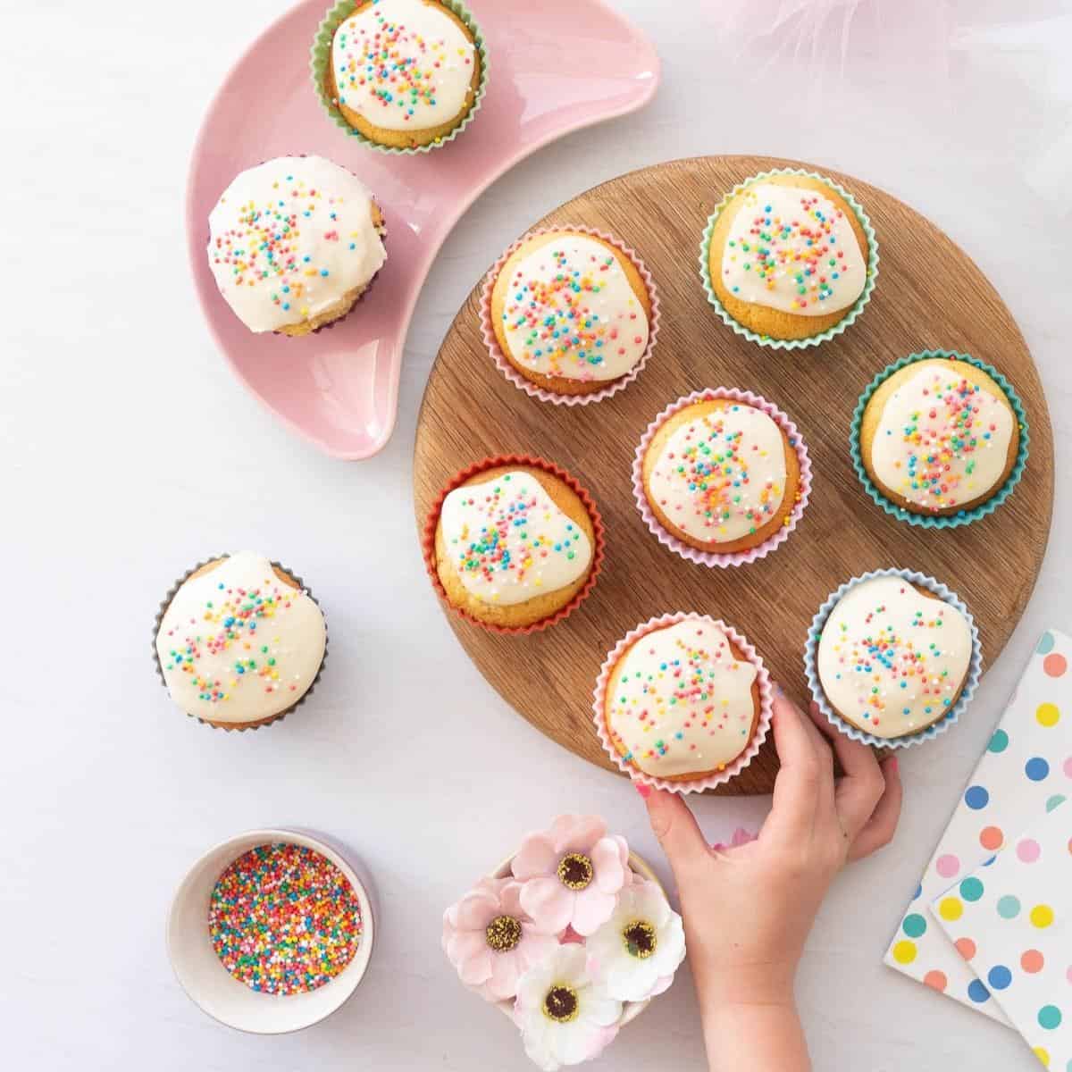 A child's hand reaching for cupcakes decorated with white frosting and sprinkles on a wooden chopping board.