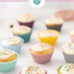 Cupcakes in pastel coloured cupcake cases on a white bench top sprinkled with rainbow sprinkles, text overlay for pinterest.