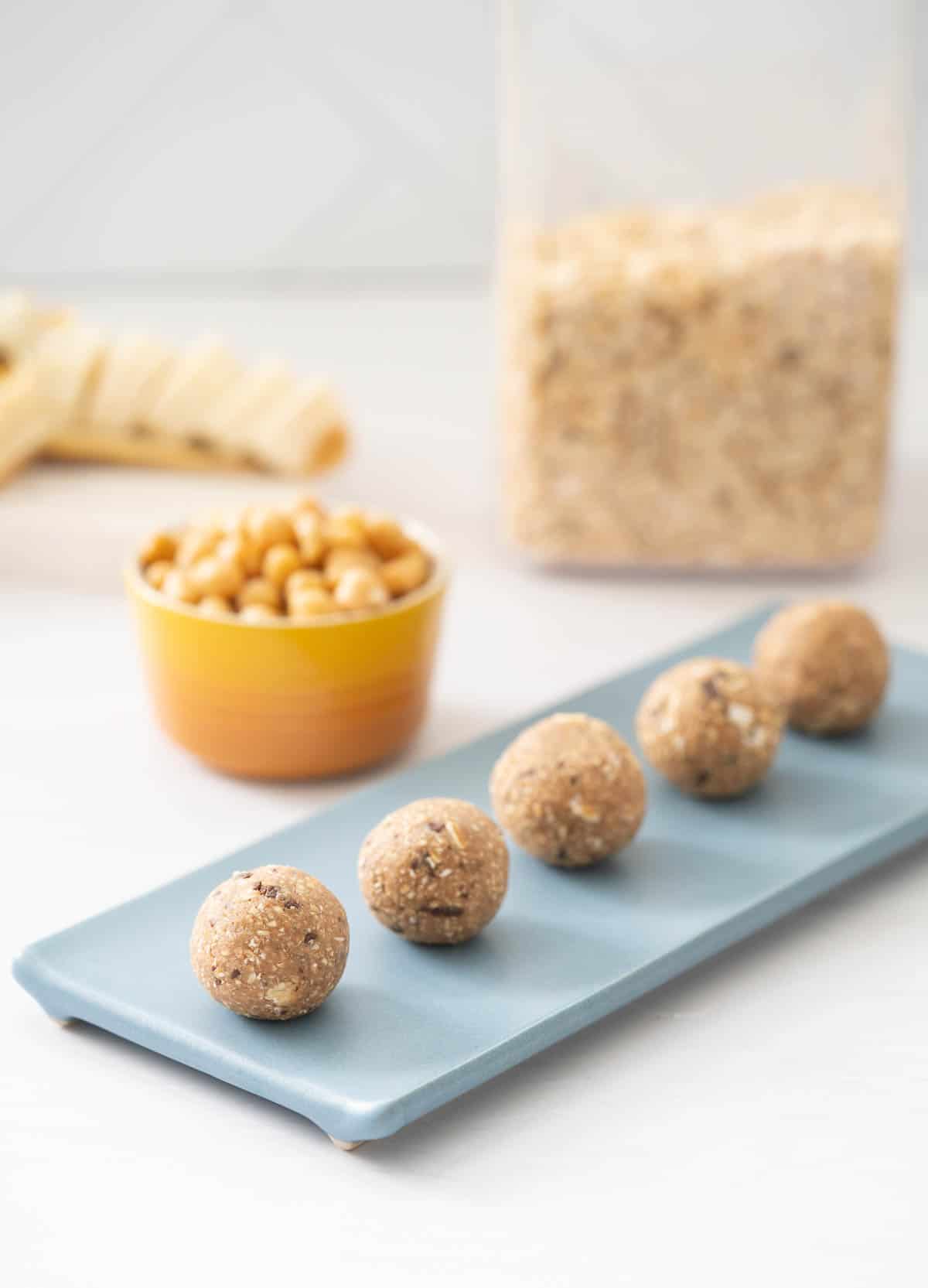 Bliss balls lined up on a small blue plate with a bowl of chickpeas and a large container of rolled oats in the background.