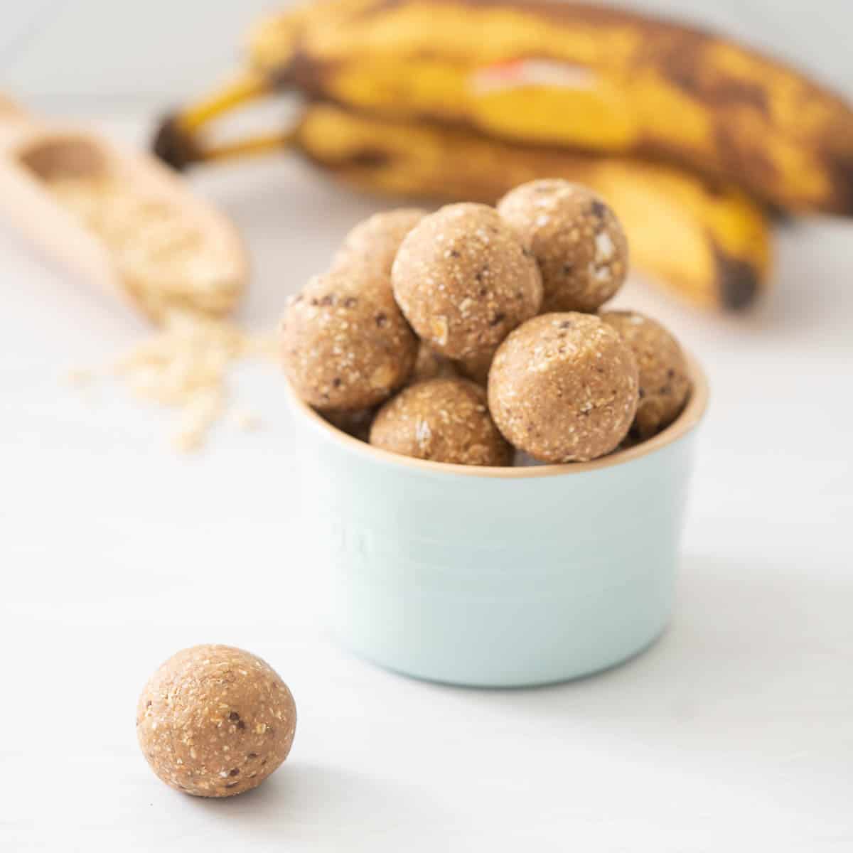 A banana flavoured energy bite sitting in-front of a blue ceramic bowl of more banana bread balls, ripe bananas and rolled oats in the background.