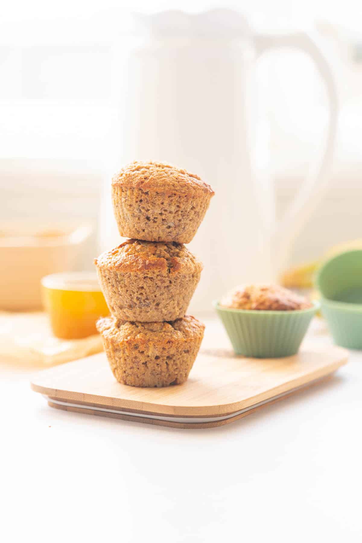 A tower of three bran muffins on a wooden board sitting in front of a white jug.