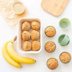 A box packed with six banana bran muffins on a bench top next to banans, a bowl of wheat bran and four more muffins in reusable muffin cups.