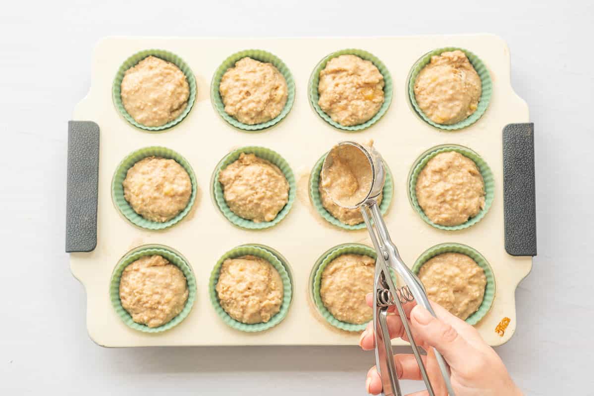 Muffin batter being scooped into a green silicone lined muffin tray. 