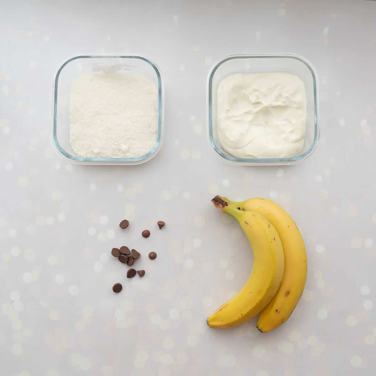 Container of yoghurt, container of desiccated coconut, two bananas and chocolate drops laid out on a bench top.