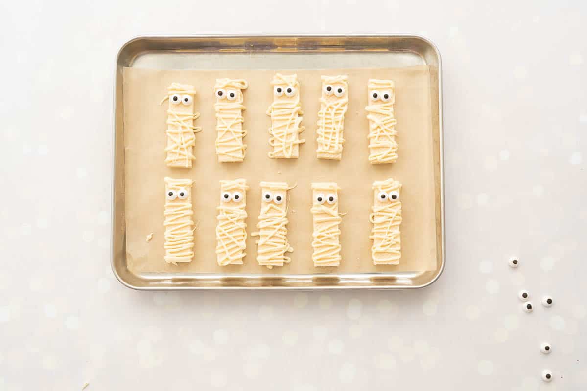 Vanilla wafer biscuits on a lined tray decorated with white chocolate drizzle and candy eyes to look like mummies.