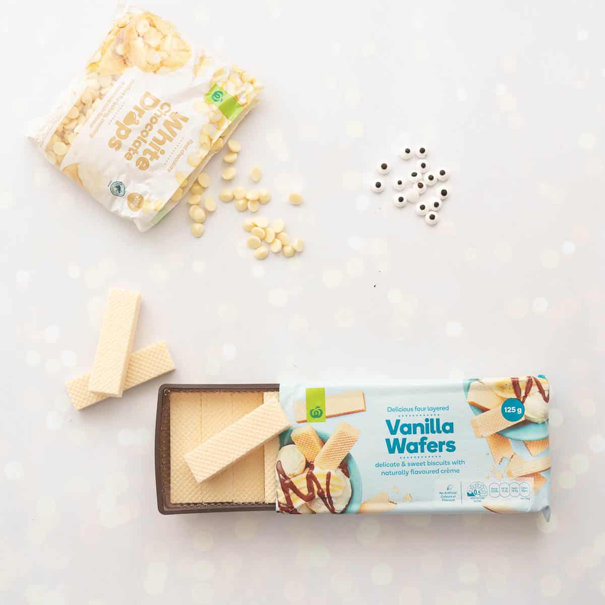 A packet of white chocolate drops, vanilla wafers and candy eyes laid out on a bench top.