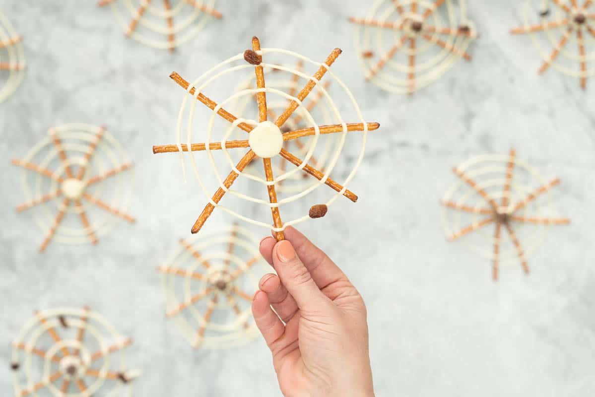 A spider web made from pretzel sticks and white chocolate, and decorated with raisins to look like flies being held up.