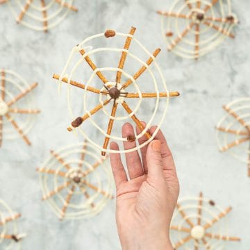 A spider web made from pretzel sticks and white chocolate, and decorated with a milk chocolate spider in the centre being held up.