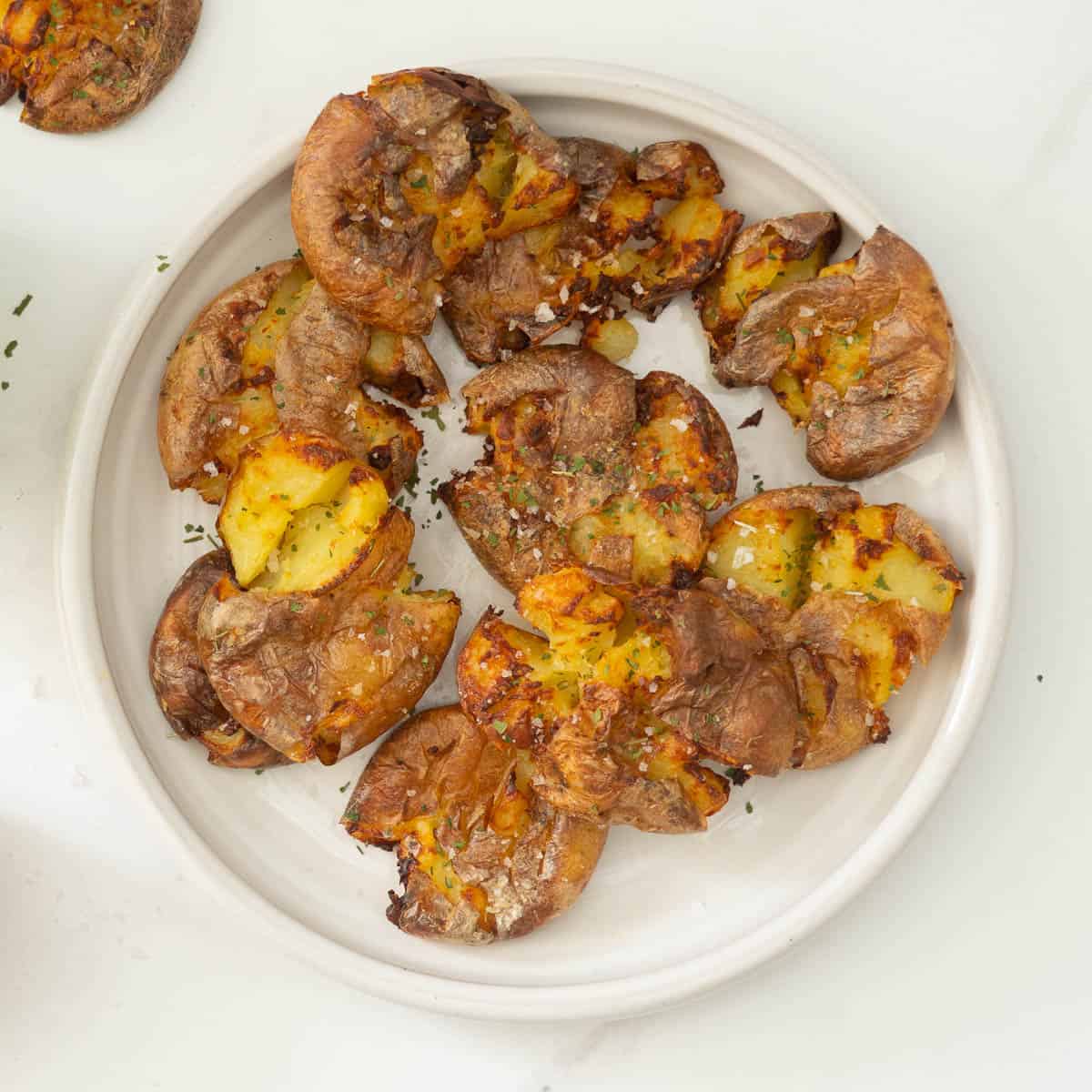 A white ceramic plate of golden smashed potatoes sprinkled with salt and parsley flakes.