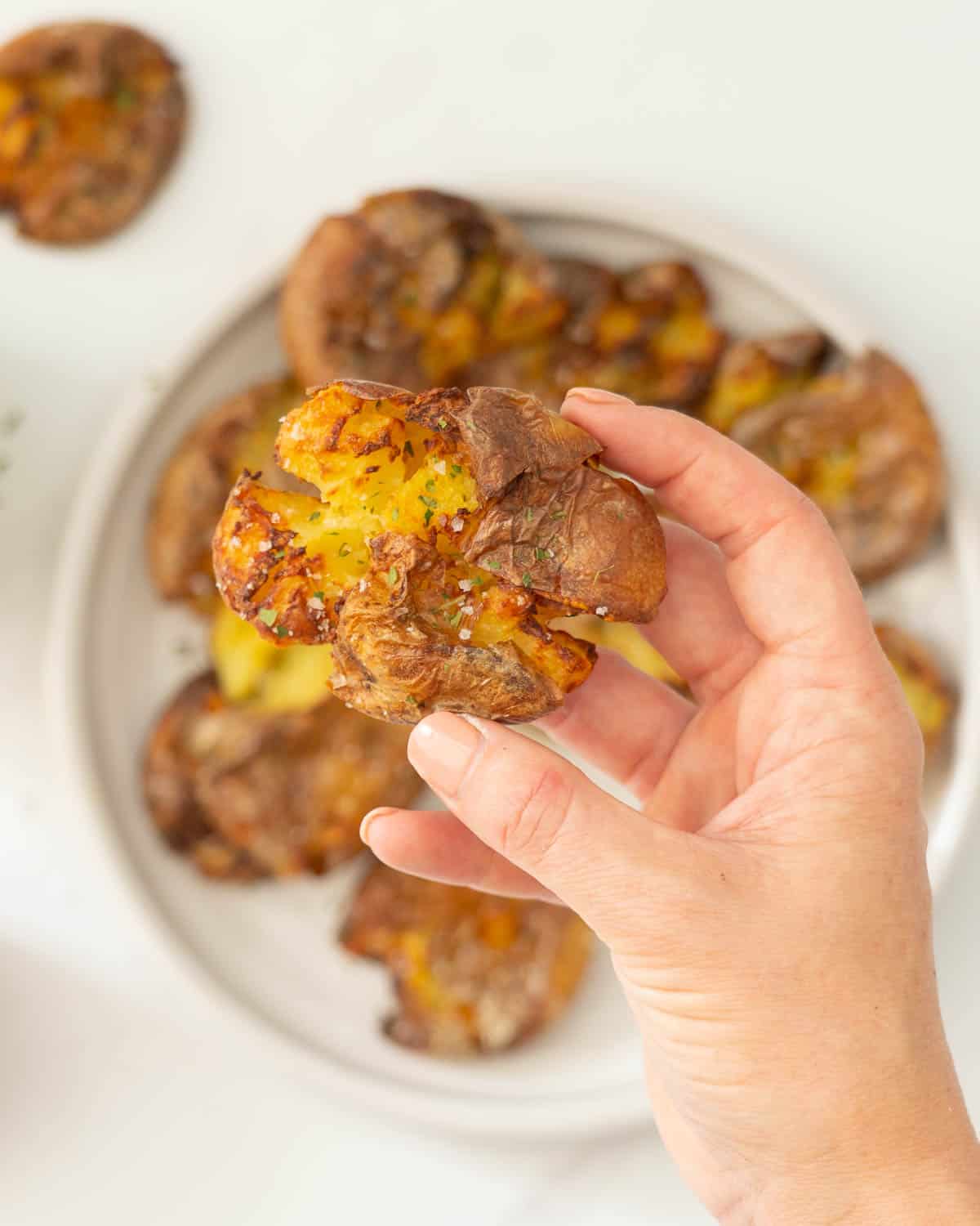 A golden brown smashed potato with skin on being held above a plate of smashed potatoes.