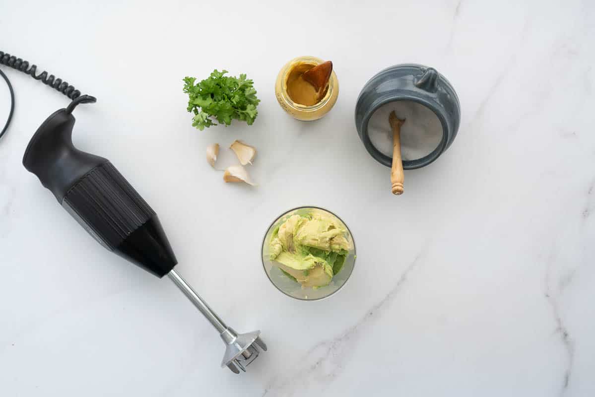 A emulsion blender, parsley. mustard, salt and garlic cloves lying on a bench top next to a blender cup full of avocado.