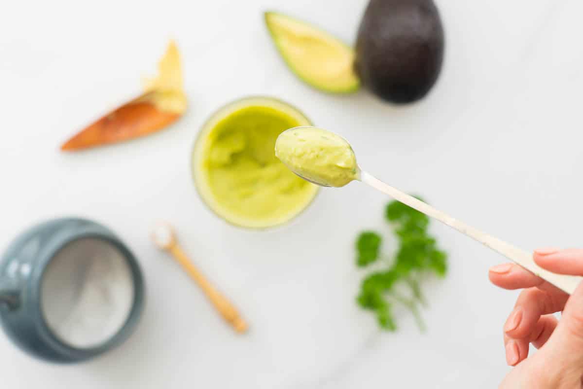 A spoonful of smooth avocado aioli being held above a blender cup of more avocado aioli.