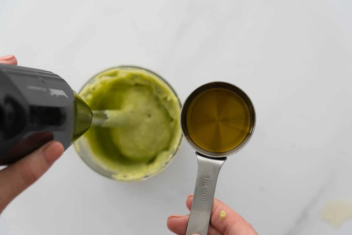 A measuring cup of oil being poured into a blender of avocado purée.