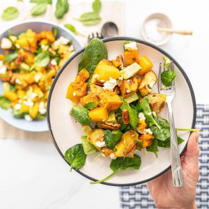 A plate of salad containing sweet potato, peaches, pecans, feta, avocado and spinach leaves being held above a dining table.