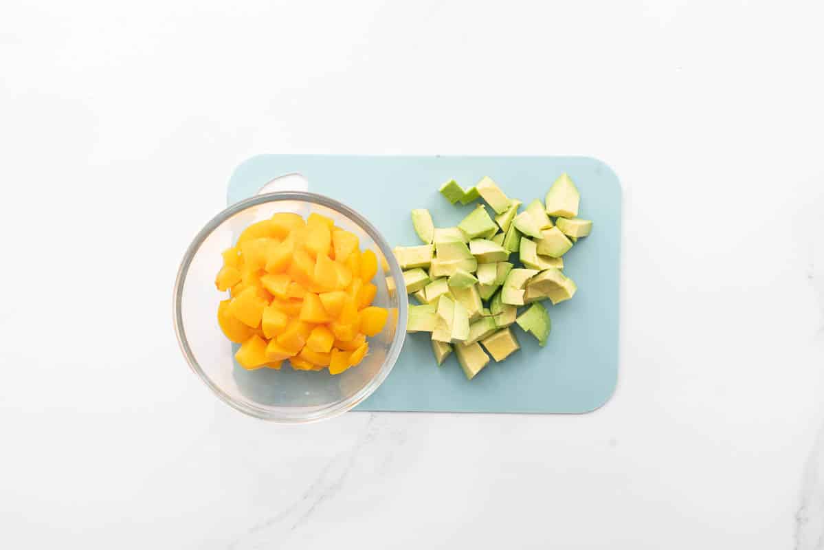 Cubes of avocado on a light blue chopping board next to a small glass bowl of cubed peaches.
