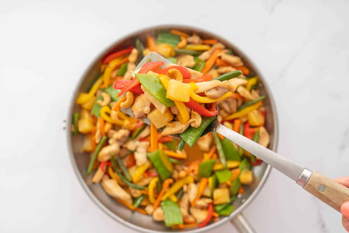 A spoonful of pineapple chicken stir-fry being scooped from a large pan of stir fry.