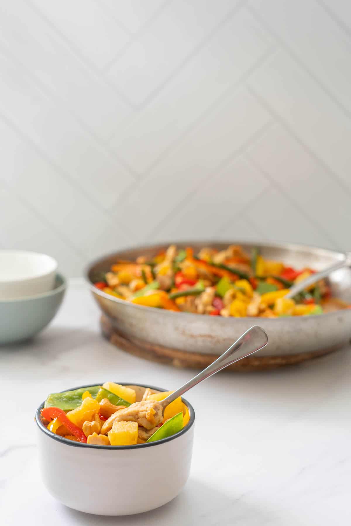 A small bowl of colourful chicken and vegetable stir fry sitting in front of a fry pan of more stir fry.