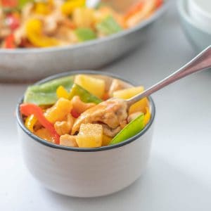 A small black rimmed white bowl filled with a colourful chicken vegetable stir fry, pieces of pineapple and cashew nuts also visible.