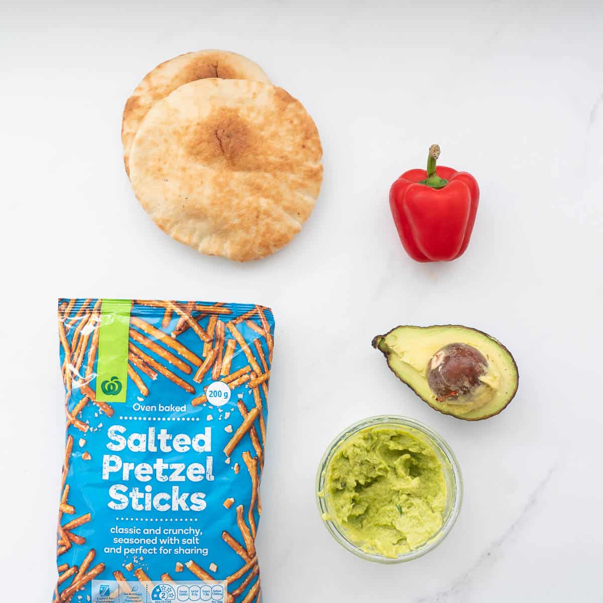 Two pita breads, a bag of pretzel sticks, a red capsiucm and half an avocado lying on a marble bench top.