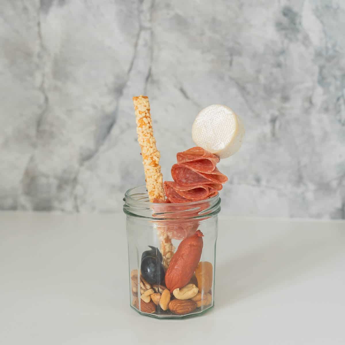 A small jar, one quarter filled with nuts and dried apricots, a bread stick and cheese on a skewer poking up and out of the jar.