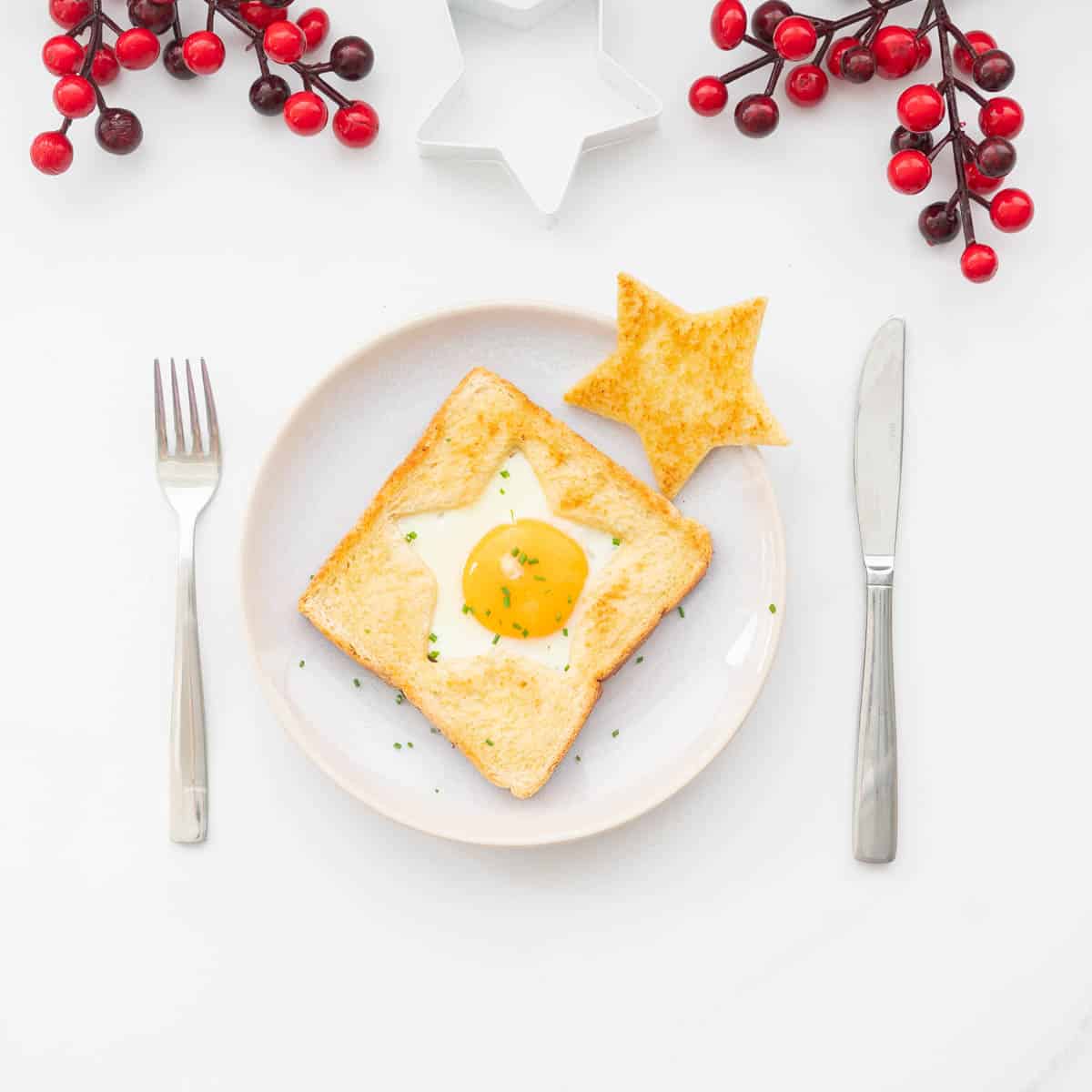 An egg cooked into the star shaped hole of a piece of toast on a plate with a golden star piece of toast to the side, red Christmas decoration around the plate.
