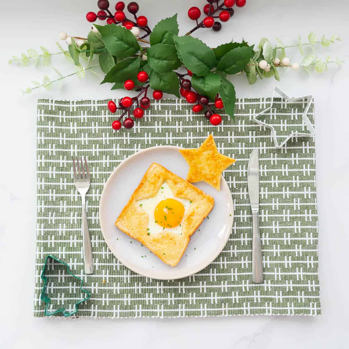 An egg cooked into the star shaped hole of a piece of toast on a plate with a golden star piece of toast to the side, sitting on.a green patterned placemat with a white green and red christmas decoration centrepiece above the placemat.