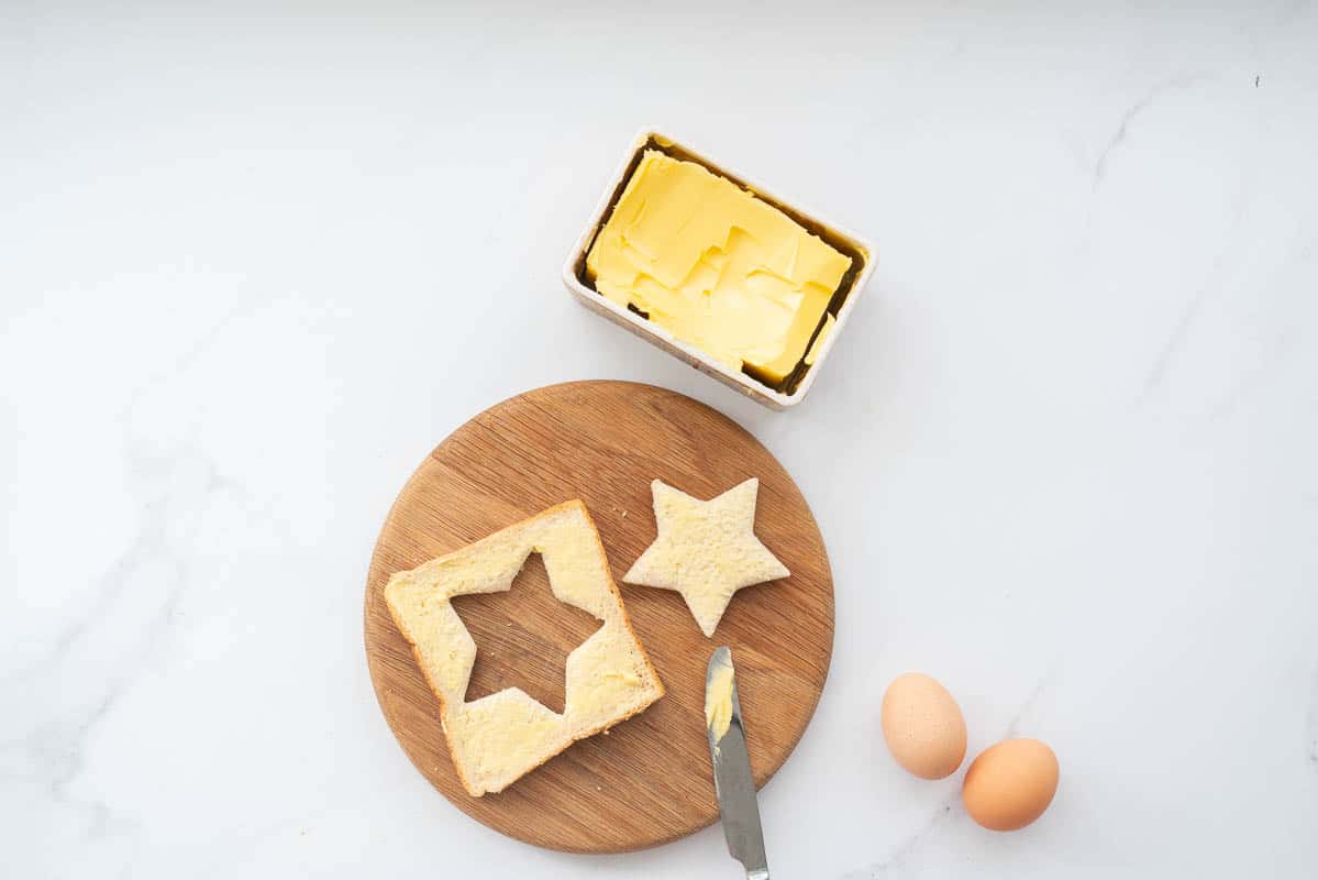 A slice of buttered bread with a star cut out of the centre on a round wooden chopping board.