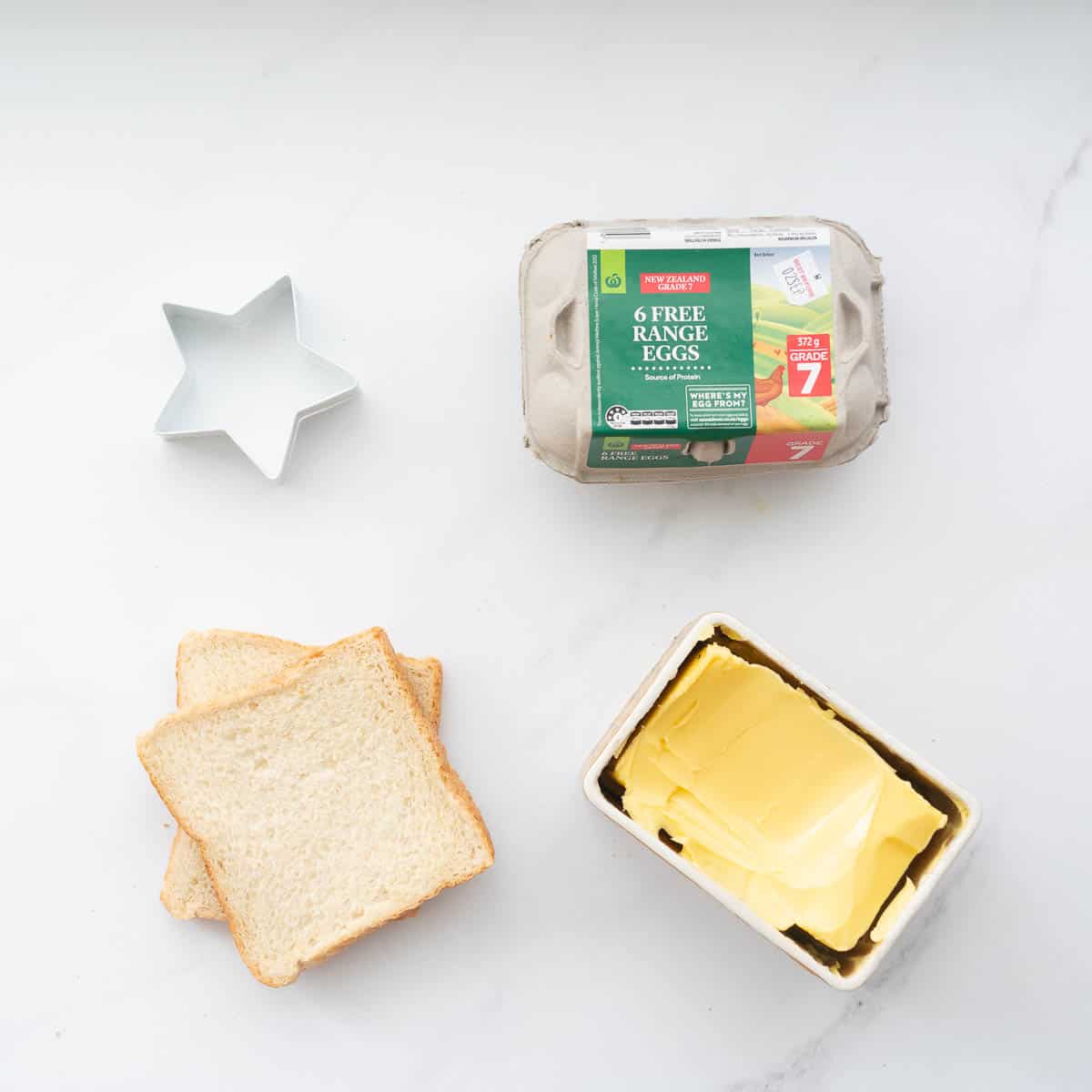 A christmas cookie cutter, 2 slices of bread, a carton of eggs and a block of butter in a dish on a bench top.