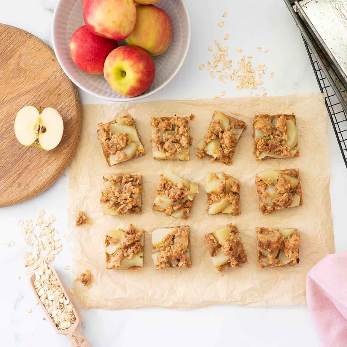 Twelve pices of apple slice on a sheet of brown parchment paper on a bench top scattered with rolled oats and apples.